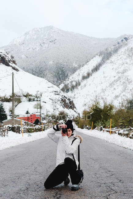 Mujer en chaqueta blanca de invierno con capucha y pantalones negros tomando fotos con la cámara mientras está de pie en medio de la carretera de asfalto - foto de stock