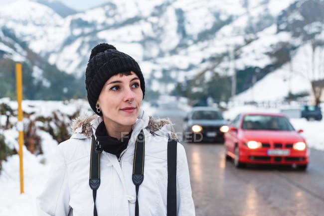 Donna a piedi in mezzo alla strada asfaltata tra montagne innevate con auto sullo sfondo — Foto stock