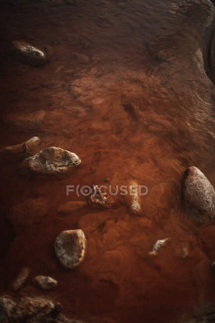 De arriba ondulante agua marrón que fluye en poco profundo con piedras hacia el acantilado en terreno rocoso - foto de stock