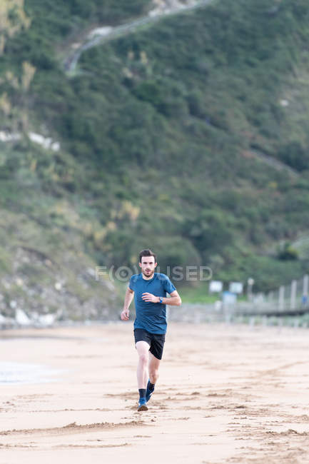 Bearded male athlete in active wear running during empty sandy beach with green mountains on blurred background — Stock Photo