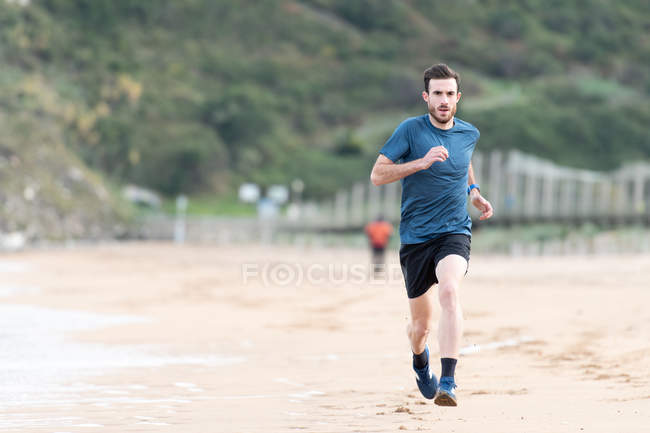 Atleta masculino barbudo en uso activo corriendo durante la playa de arena vacía con montañas verdes sobre fondo borroso - foto de stock