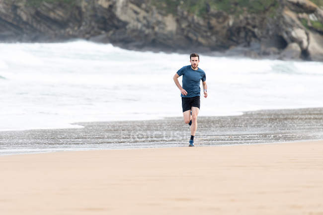 Atleta masculino barbudo em desgaste ativo correndo durante praia vazia de areia com montanhas verdes em fundo embaçado — Fotografia de Stock