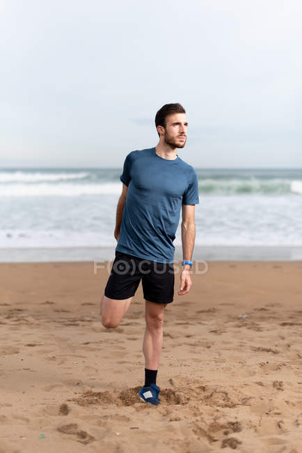 Homem desportivo no desgaste ativo esticando as pernas para correr no litoral arenoso vazio e olhando para longe — Fotografia de Stock