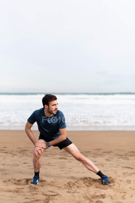 Gimnasta masculino con ropa deportiva que estira las piernas y mira hacia otro lado en la playa de arena vacía con el mar azul y el cielo sobre fondo borroso - foto de stock