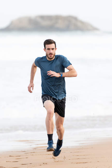 Atleta maschio barbuto in usura attiva che corre durante la spiaggia sabbiosa vuota con montagne verdi su sfondo sfocato — Foto stock