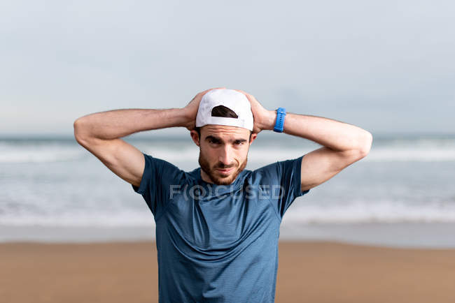 Sportsman en t-shirt bleu avec les mains derrière la tête sur capuchon blanc en regardant la caméra avec bord de mer sablonneux vide sur fond flou — Photo de stock