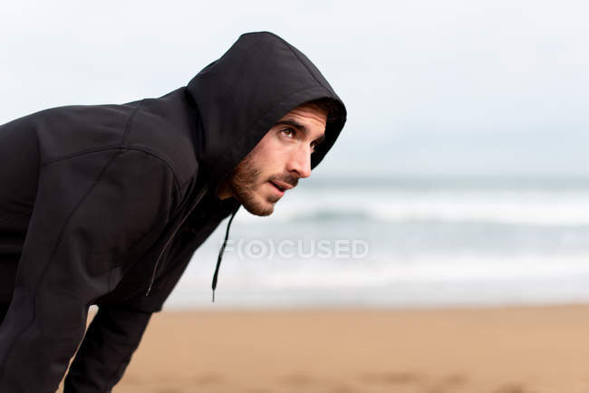 Ginasta macho barbudo com capuz preto de pé com as mãos nos joelhos e olhando para longe com praia de areia no fundo borrado — Fotografia de Stock