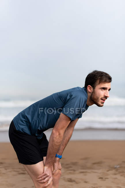 Vista lateral del hombre deportivo barbudo en desgaste activo tomando un descanso después de una larga carrera en la playa vacía de arena y mirando hacia otro lado - foto de stock