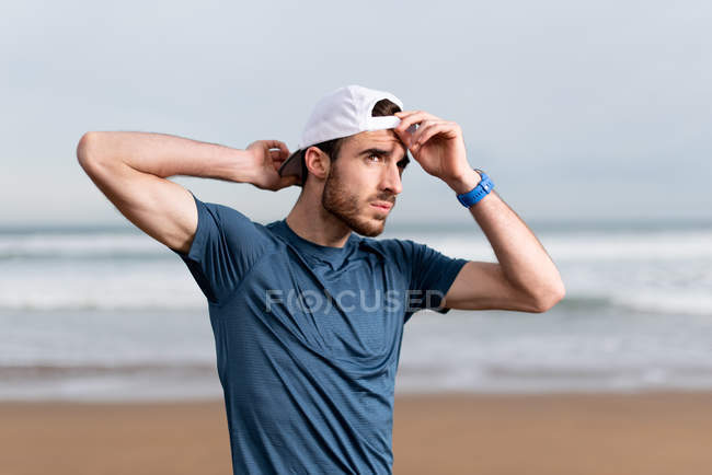 Sportler im blauen T-Shirt mit den Händen hinter dem Kopf auf der weißen Mütze, der mit leerem Sandstrand auf verschwommenem Hintergrund wegschaut — Stockfoto