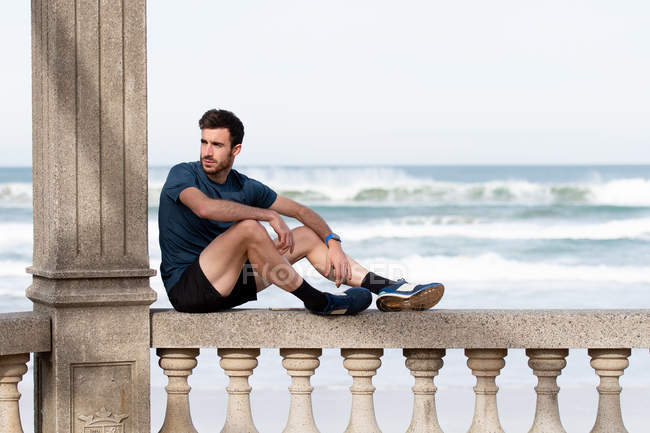 Side view of bearded male gymnast in active wear resting on rocked fence after train and looking away with sea and sky on blurred background — Stock Photo