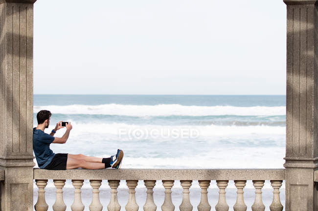 Vista lateral del hombre sin rostro en desgaste activo sentado en la valla mecedora y apoyado en la columna mientras toma una foto del mar azul en el teléfono móvil - foto de stock