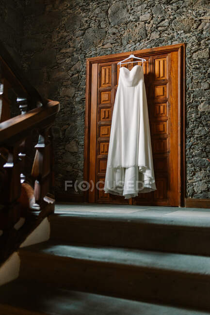Robe de mariée blanche suspendue à la porte en bois sur escalier dans la maison avec mur en pierre — Photo de stock