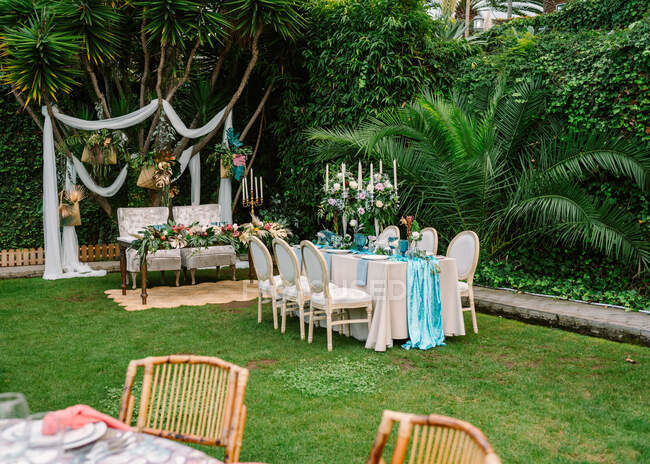 Decoración para la ceremonia de boda al aire libre con mesa de cena en colores azul y blanco y ajuste de arco de boda en el jardín con plantas tropicales verdes - foto de stock
