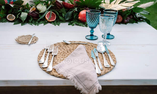 Білий дерев'яний стіл без тканини, прикрашений аранжуванням з фруктами та квітами і подається з столовими приборами та лляною серветкою на плетених серветках та блакитних окулярах — стокове фото