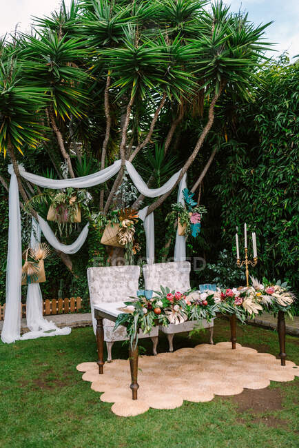 Set für die Trauung mit frisch Vermählten Tisch mit Blumenarrangements dekoriert und auf Teppich gegen grünen tropischen Baum mit weißem Hochzeitsbogen gelegt — Stockfoto