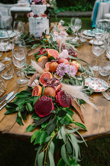 Beautiful garland made from ripe cut tropical fruits and green branches with flowers and decorative plants setting on round wooden wedding table served with plates and glasses — Stock Photo