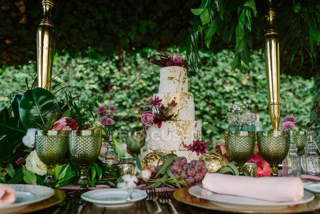 White and gold tiered cake with flowers placed on wedding table with plate and glasses decorated with fruits and flowers against green trees — Stock Photo