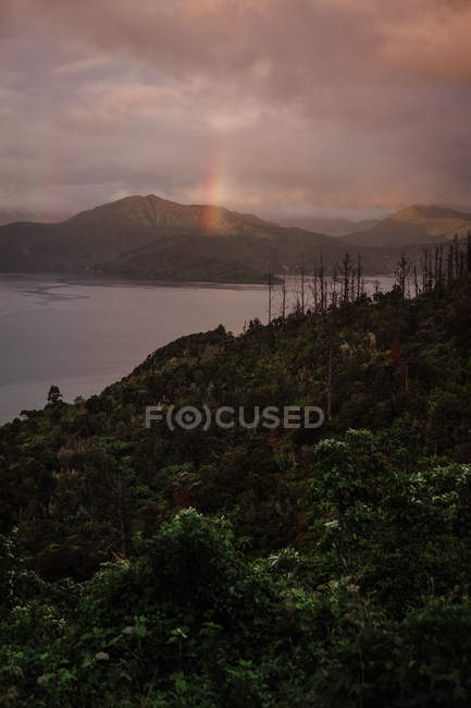 Spectacular landscape with bay shore surrounded by green hills and rainbow over mountain on cloudy day in New Zealand — Stock Photo