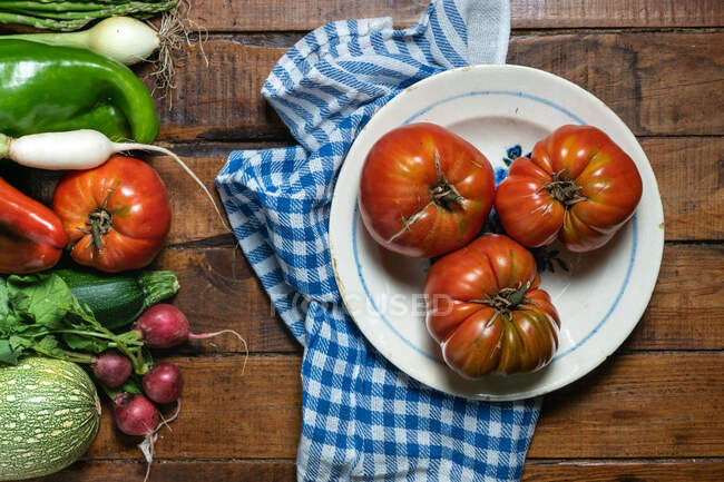 Вид сверху на старую тарелку с большими красными помидорами, расставленными на голубой и белой салфетках на дощатом деревянном столе со смешанными свежими овощами, отложенными в сторону — стоковое фото