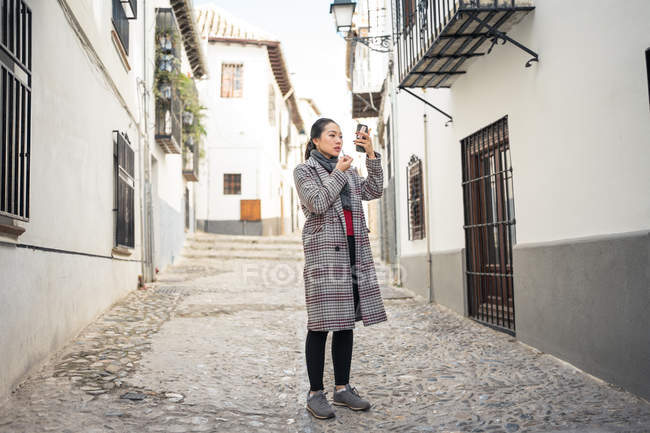 Young Asian female traveler in stylish casual coat refreshing makeup while looking in screen of mobile phone among street — Stock Photo