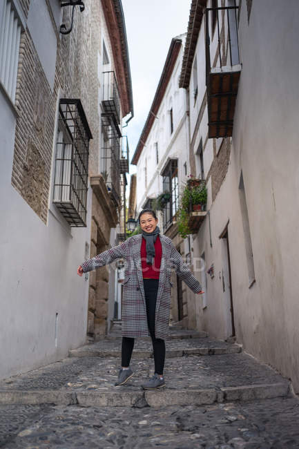 Снизу веселая женщина в повседневной одежде улыбается и стоит, поднимая руки и глядя в камеру среди переулка Альбайсин в Гранаде — стоковое фото