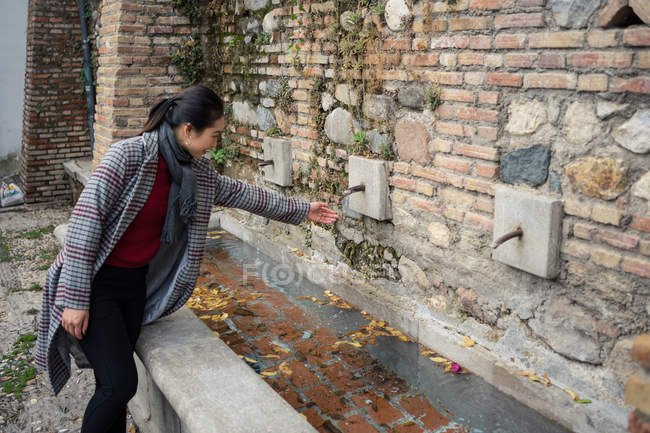 Vue latérale d'une curieuse femme aux cheveux noirs vêtue de vêtements décontractés essayant de se laver les mains dans la fontaine d'un vieux bâtiment rocailleux dans la rue — Photo de stock