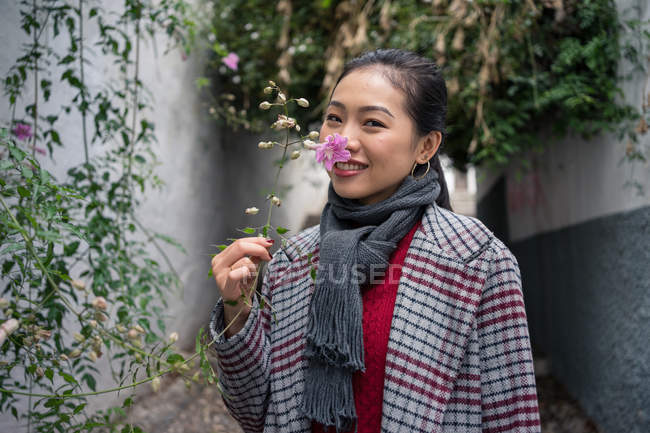 Молодая женщина в повседневной одежде прикасается к зеленым растениям, нюхает цветы и улыбается на раскачивающемся переулке — стоковое фото
