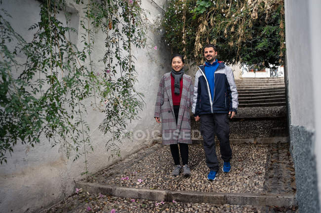 Содержите полиэтническую пару в повседневной одежде, держась за руки во время прогулки по раскачивающимся старым улицам с зелеными растениями — стоковое фото