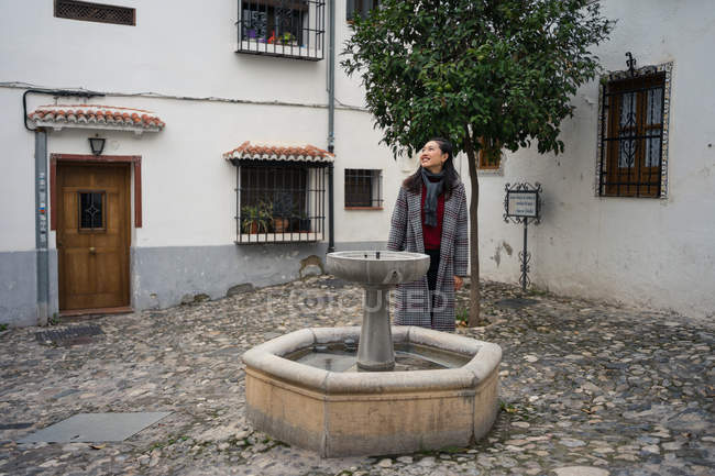 Femminile turista in casuale cappotto in piedi su piazza ciottoli con piccola fontana tra vecchi edifici — Foto stock