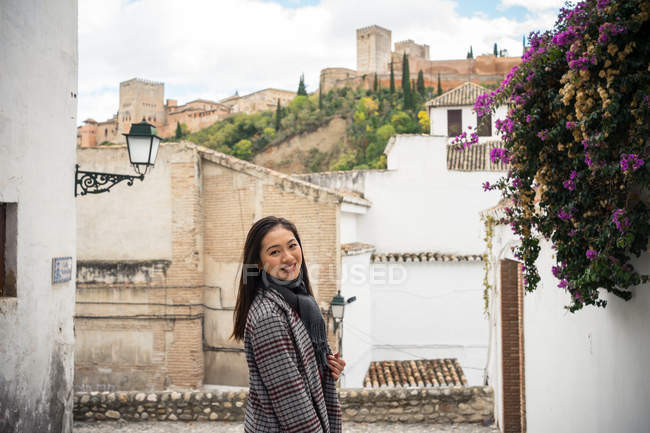 Vista lateral do turista feminino asiático satisfeito em casaco elegante olhando na câmera com edifícios antigos fodidos e belo castelo — Fotografia de Stock