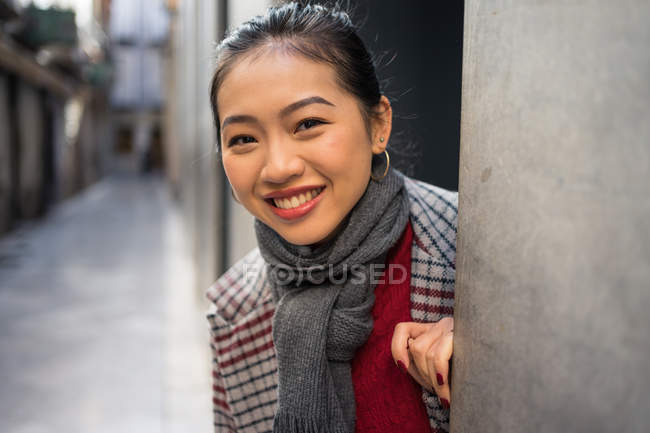 Захоплені радістю азіатська жінка у звичайному пальто, що вдивляється з стіни сучасного будинку і дивиться в камеру в Альбайцині (Гранада, Іспанія). — стокове фото