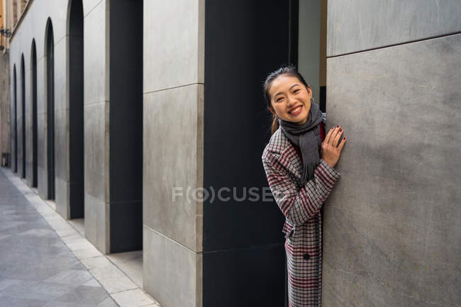 Mulher asiática muito feliz em casaco casual espiando da parede do edifício moderno e olhando na câmera em Albaicin em Granada, Espanha — Fotografia de Stock