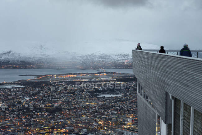 Back view of faceless travelers standing on roof of contemporary building and contemplating amazing view of evening city located along coastline in valley against snowy highland — Stock Photo