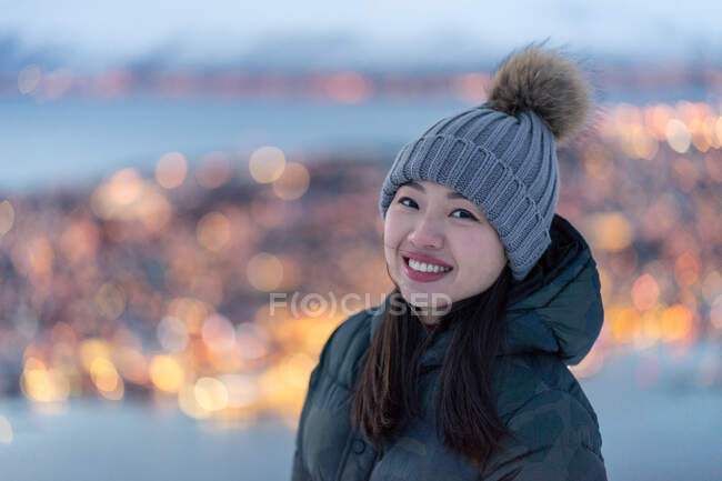 Aufgeregte junge Frau in khakifarbener Daunenjacke und grauem, warmem Hut, die in die Kamera schaut und am Abend erstaunliche Winterbilder von der Stadt an der Küste betrachtet — Stockfoto