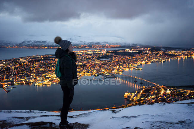 Excited young female in khaki down jacket and gray warm hat looking away and contemplating amazing winter view of city located on coast in evening — Stock Photo