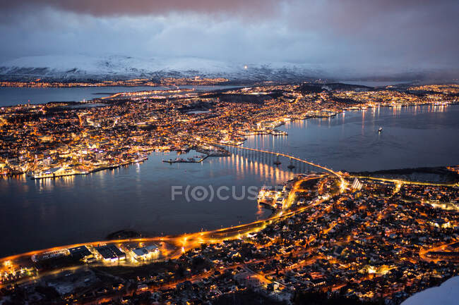 Magnificent scenery of city with golden lights located on island and shores of strait against foggy hills covered with snow under lush clouds in winter night — Stock Photo