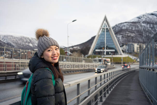 Привлекательная взрослая азиатская женщина в теплой одежде с рюкзаком, улыбающимся в камеру, стоя на улице перед размытым внешним видом удивительной церкви в форме треугольника и снежных холмов в Норвегии — стоковое фото