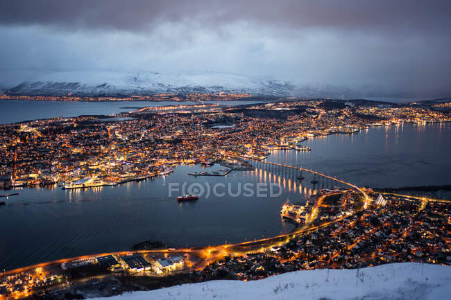 Magnifico scenario della città con luci dorate situate sull'isola e sponde dello stretto contro colline nebbiose coperte di neve sotto lussureggianti nuvole nella notte d'inverno — Foto stock