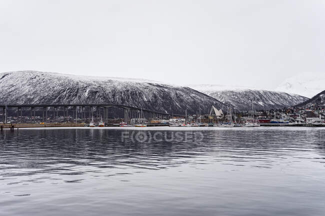 Desde arriba de la bahía tranquila con la ciudad en la costa ordenada poderosa montaña nevada con cielo nublado en el fondo en Tromso en Noruega - foto de stock