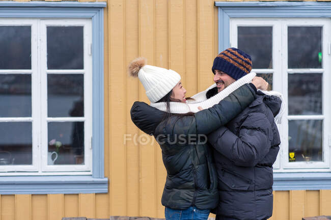 Multiétnica feliz pareja de turistas en ropa de abrigo abrazando cerca de casa rural - foto de stock