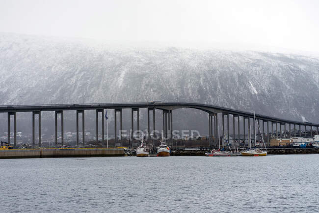 Современный мост и лодки на спокойном море с большой снежной горой на заднем плане в Норвегии — стоковое фото