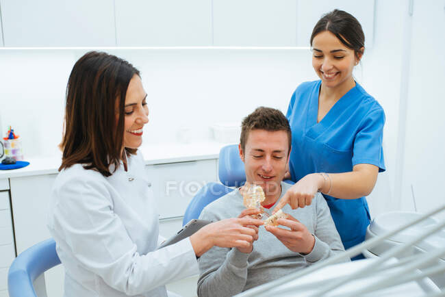 Odontólogo sonriente y asistente mostrando estructura de dientes en prótesis al cliente en silla - foto de stock