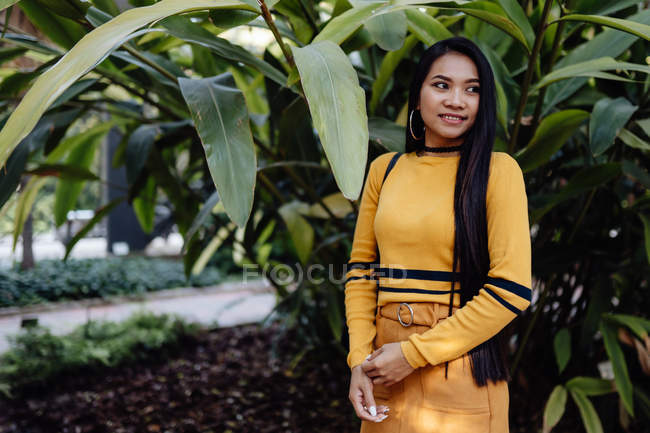 Vista lateral de la mujer asiática de moda con pelo largo y oscuro en camisa amarilla y falda corta de pie en un hermoso jardín y mirando hacia otro lado - foto de stock