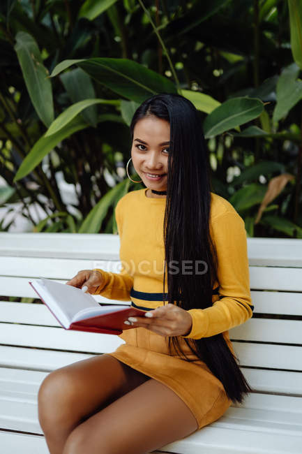 Brünette asiatische Frau liest Buch in rotem Einband sitzt auf weißer Bank im Park — Stockfoto