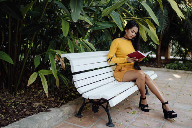 Vista laterale di bruna donna asiatica che legge il libro in copertina rossa seduta su una panchina bianca nel parco — Foto stock