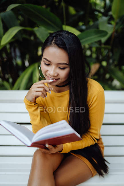 Side view of brunette Asian woman reading book in red cover sitting on white bench in park — Stock Photo