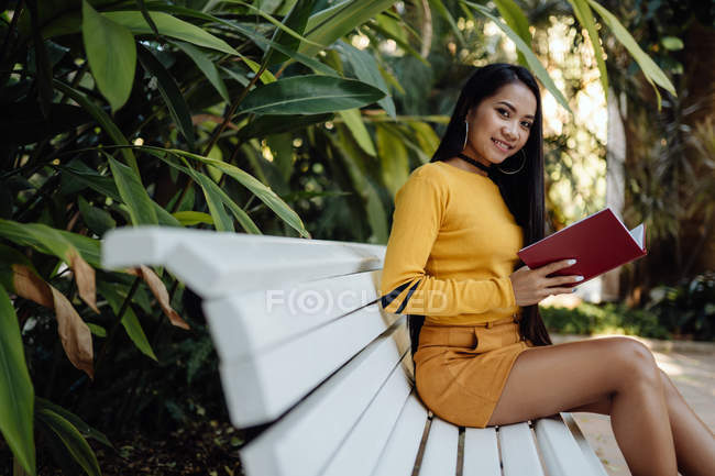 Вид сбоку брюнетки азиатской женщины, читающей книгу в красной обложке, сидящей на белой скамейке в парке — стоковое фото
