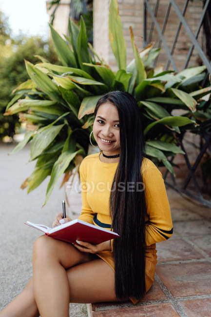Asiatico donna lettura libro in rosso copertina mentre seduta in un edificio ingresso vicino verde pianta su un pot su il strada — Foto stock