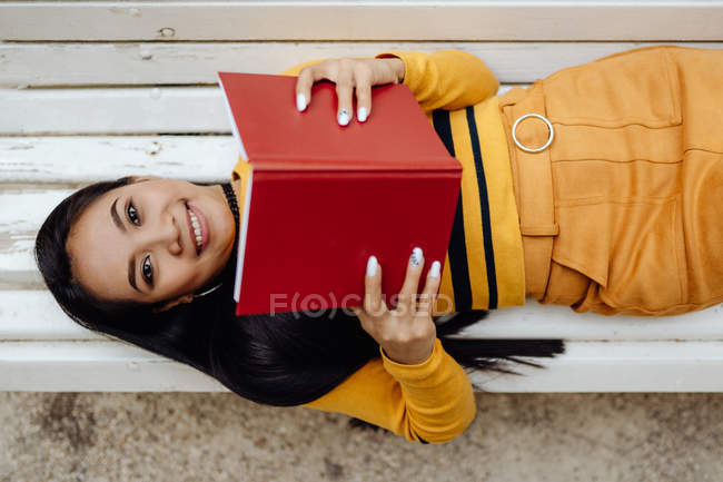 From above of brunette Asian woman reading book in red cover lying down on white bench in park — Stock Photo