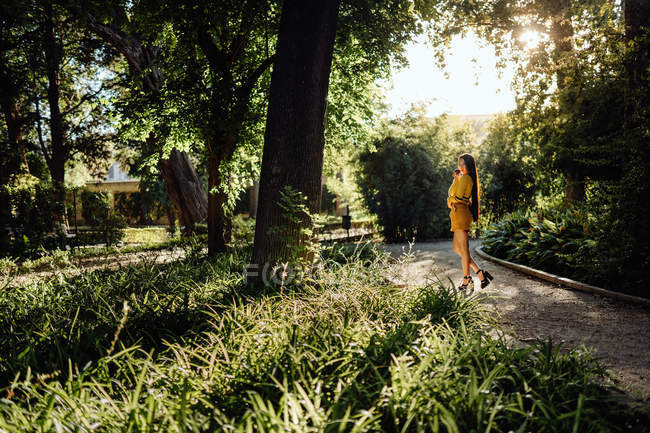 Mujer asiática con cabello largo y oscuro caminando por el camino iluminado por el sol y el prado de hierba mirando a la cámara sobre el hombro - foto de stock